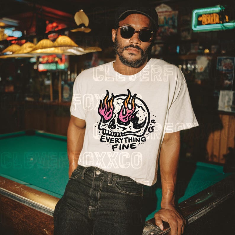 Puede incluir: Camiseta color crema con un gr&aacute;fico de calavera con llamas rosas y naranjas, y el texto "Everything Fine". La camiseta se combina con vaqueros oscuros. La imagen est&aacute; ambientada en un bar con poca luz.