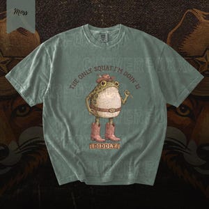 A t-shirt with a cartoon frog wearing cowboy boots and a hat, along with the text "The only squat I'm done is Diddly" printed on it.