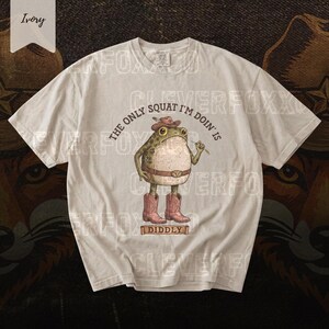 A white t-shirt with a cartoon frog wearing cowboy boots and a hat, along with the text "The only squat I'm done is" and "Diddyly" printed on it.