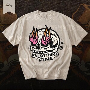 Puede incluir: Camiseta color marfil con un dise&ntilde;o de calavera con llamas rosas y naranjas. El texto "EVERYTHING FINE" est&aacute; impreso en negro. La camiseta de manga corta tiene cuello redondo.