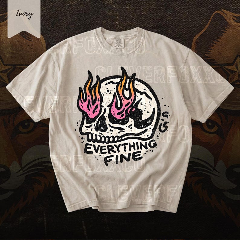 Puede incluir: Camiseta color marfil con un dise&ntilde;o de calavera con llamas rosas y naranjas. El texto "EVERYTHING FINE" est&aacute; impreso en negro. La camiseta de manga corta tiene cuello redondo.