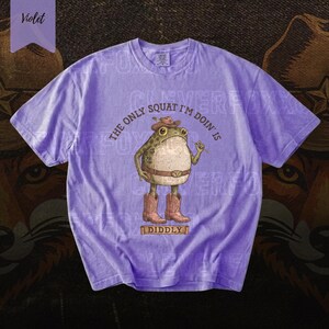 A purple t-shirt with a cartoon frog wearing a cowboy hat and boots, along with the text "The only squat I'm doing is I didlyy" printed on it.