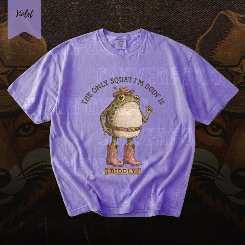 A purple t-shirt with a cartoon frog wearing a cowboy hat and boots, along with the text "The only squat I'm doing is I didlyy" printed on it.