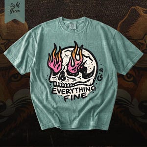 Puede incluir: Camiseta verde claro con un gr&aacute;fico de calavera y llamas rosas y naranjas. El texto "EVERYTHING FINE" est&aacute; debajo de la calavera. La camiseta es de color verde claro.