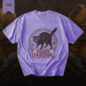 A purple t-shirt with a graphic design featuring a cat and the text "FIGHT FASCISM" printed on it.