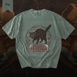 A gray t-shirt with a graphic design featuring a cat and the text "FIGHT FASCISM" printed on it.