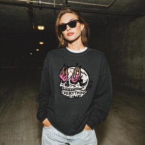 Puede incluir: Sudadera gris oscuro con un gr&aacute;fico de calavera con llamas rosas y amarillas y el texto "Everything Fine". La sudadera tiene cuello redondo y mangas largas. La persona lleva gafas de sol negras.