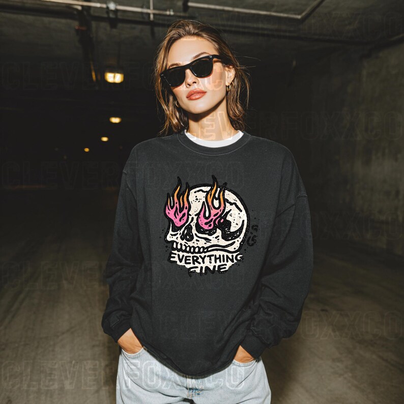 Puede incluir: Sudadera gris oscuro con un gr&aacute;fico de calavera con llamas rosas y amarillas y el texto "Everything Fine". La sudadera tiene cuello redondo y mangas largas. La persona lleva gafas de sol negras.