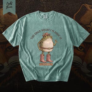 A light blue t-shirt with a cartoon frog wearing cowboy boots and a hat, along with the text "The only squat I'm done is I didlyy" printed on it.
