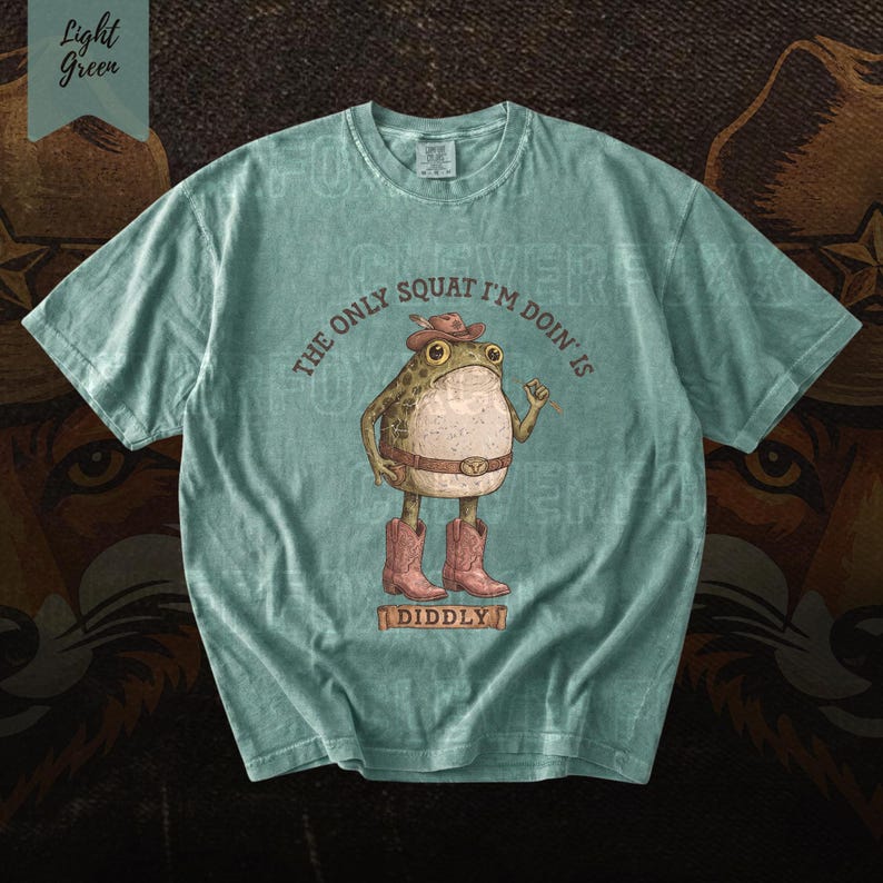 A light blue t-shirt with a cartoon frog wearing cowboy boots and a hat, along with the text "The only squat I'm done is I didlyy" printed on it.