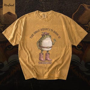 A yellow t-shirt with a cartoon frog wearing a cowboy hat and boots, along with the text "The only squat I'm done is I didlyy" printed on it.