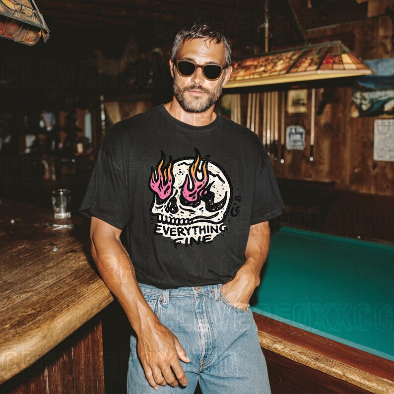 Puede incluir: Camiseta negra con un dise&ntilde;o de calavera y el texto "Everything Fine". La calavera tiene llamas rosas y amarillas. La camiseta se combina con vaqueros azules.