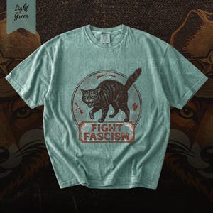 A light blue t-shirt with a graphic design featuring a cat and the text "FIGHT FASCISM" printed on it.