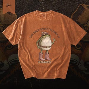a t-shirt with a cartoon frog wearing boots and a hat, along with the text "The only squat I'm doing is Diddly" printed on it.