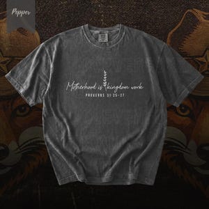 Puede incluir: Camiseta gris oscuro con el texto blanco "Motherhood is kingdom work" y "Proverbios 31:25-27". La camiseta tiene cuello redondo y mangas cortas. La palabra "Pepper" está en la esquina superior izquierda.