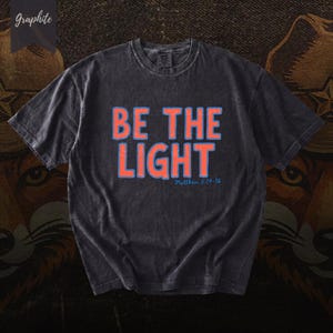 Puede incluir: Camiseta gris grafito con las palabras "BE THE LIGHT" en letras naranja con contorno azul. El texto "Matthew 5:14-16" está debajo. La camiseta tiene un aspecto vintage y desgastado.