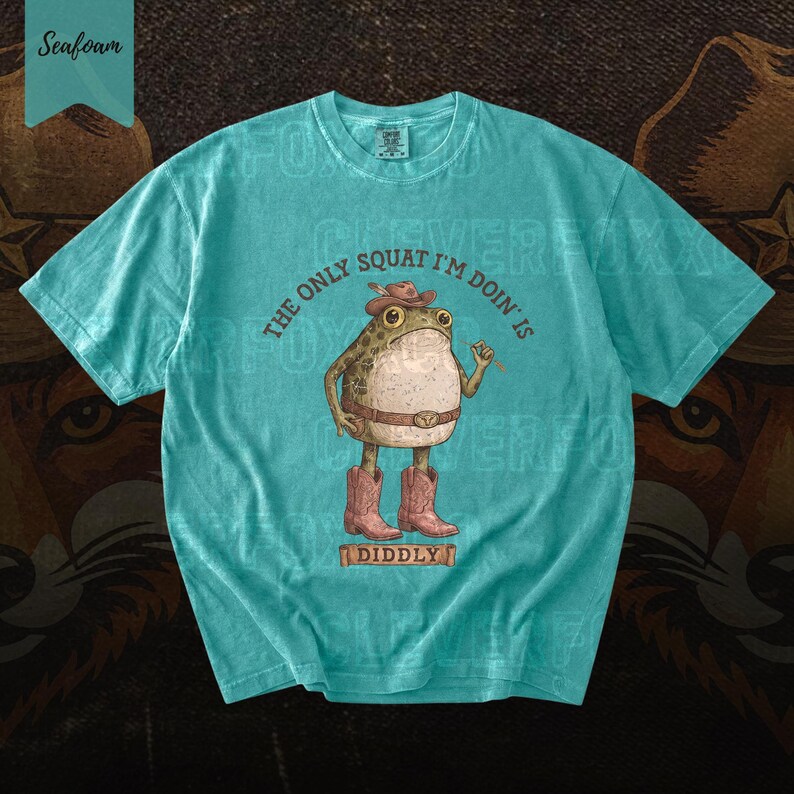 A turquoise t-shirt with a cartoon frog wearing cowboy boots and a hat, along with the text "The only squat I'm doing is I didly" printed on it.