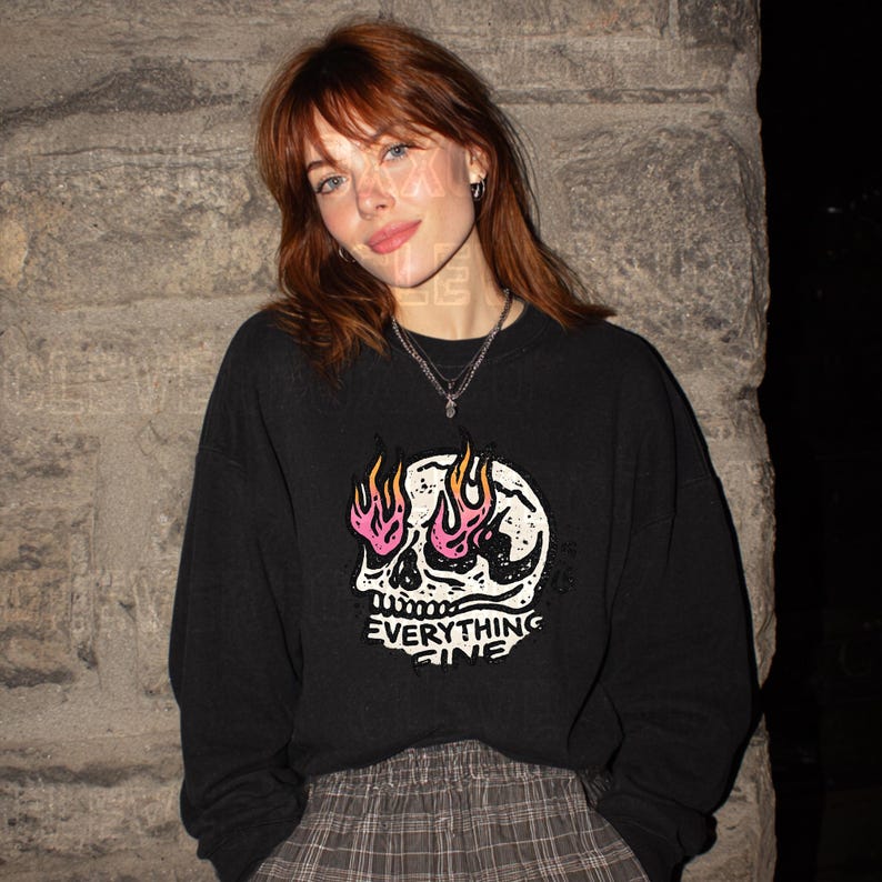 Puede incluir: Sudadera negra con un gr&aacute;fico de calavera blanca con llamas rosas y naranjas. El texto "EVERYTHING FINE" est&aacute; debajo de la calavera. La sudadera la lleva una persona pelirroja.