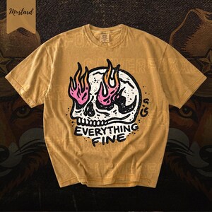 Puede incluir: Camiseta color mostaza con un dise&ntilde;o de calavera con llamas rosas y naranjas. El texto "EVERYTHING FINE" est&aacute; debajo de la calavera. La camiseta tiene mangas cortas y cuello redondo.