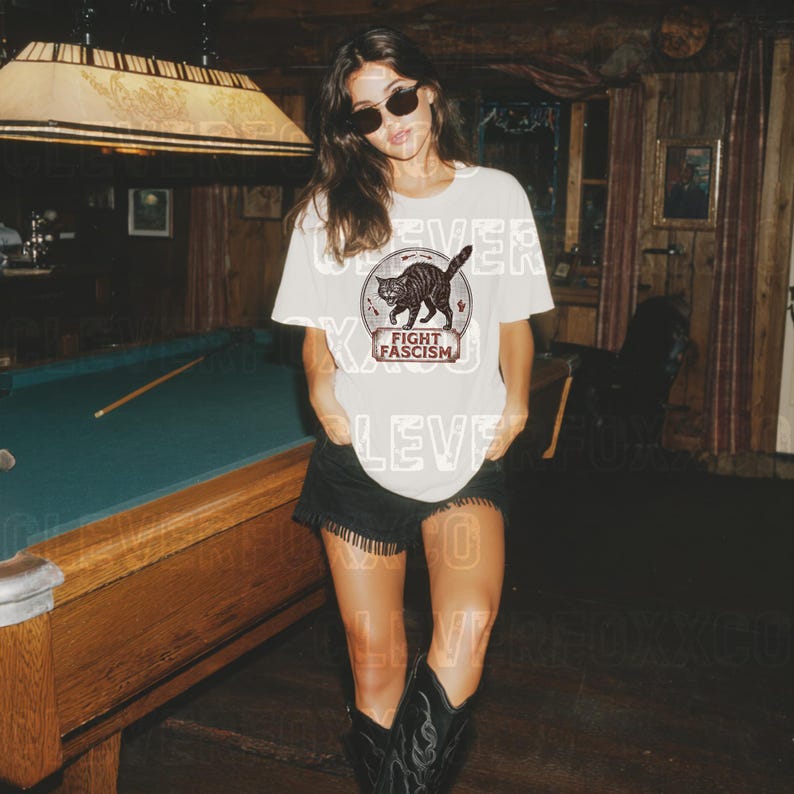 A woman wearing a white t-shirt with a graphic design and sunglasses stands in front of a pool table.