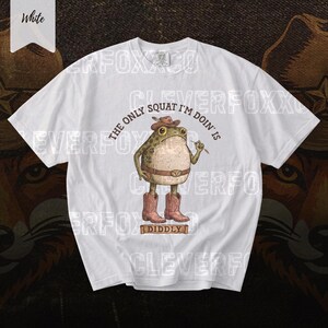 A white t-shirt with a graphic design featuring a frog character wearing cowboy boots and holding a cigarette.