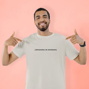 May include: A white t-shirt with the text "[SPEAKING IN SPANISH]" in black, worn by a person. The person is pointing at the shirt with both hands. The background is a solid coral color.