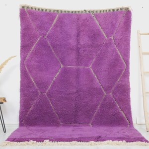 May include: A large, plush, purple rug with a geometric diamond pattern. The rug has a thick pile and a cream-colored fringe along the bottom edge. The diamond pattern is outlined in a lighter color, creating a contrasting design.