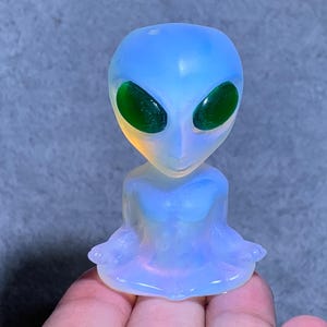 Op de afbeelding: Een doorschijnend alienbeeldje met een blauwe kop en lichaam en grote groene ovale ogen. Het beeldje heeft een glad, gepolijst oppervlak en wordt in een hand gehouden.
