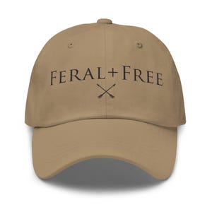 May include: A tan baseball cap with the text "FERAL + FREE" in black, with two crossed arrows. The cap has a curved brim and a button on top.