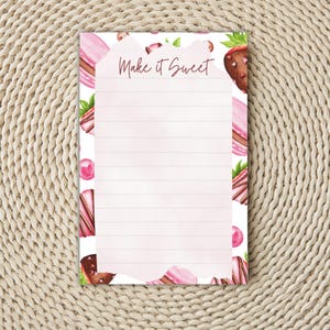 May include: A lined notepad with a watercolor design featuring pink macarons, chocolate-covered strawberries, and pink candies. The text "Make it Sweet" is written at the top in a script font. The notepad is on a woven surface.