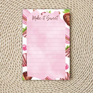 May include: A pink lined notepad with the text "Make it Sweet" in a handwritten font. The notepad is decorated with watercolor illustrations of chocolate-covered strawberries, macarons, and pink candies.