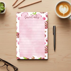 May include: A lined notepad with the words "Sweet notes" in script. The notepad has a pink watercolor wash background and is decorated with watercolor illustrations of macarons, chocolates, and cherries. The notepad is surrounded by pencils, a pen, and a cup of coffee.