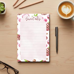 May include: A lined notepad with a watercolor design featuring pink macarons, chocolate-covered strawberries, and the text "Sweet notes." The notepad is surrounded by a pen, a cup of coffee, pencils, and a small plant.