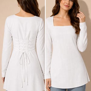 May include: A long-sleeved white tunic top featuring a square neckline and a corset-style lace-up detail on the back. The garment has a fitted bodice that flares slightly at the hem, constructed from a solid white fabric with a subtle texture.