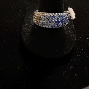May include: A wide band ring featuring a pavé setting of small round stones in a gradient color pattern. The stones transition from light champagne and white tones on the left side to various shades of blue on the right side of the metal band.