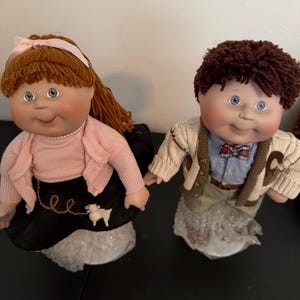 May include: Two vintage Cabbage Patch Kids dolls. The girl doll has auburn hair, a pink sweater, and a black skirt. The boy doll has brown hair and a cream-colored sweater. Both dolls have blue eyes and are holding hands.