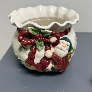 May include: A cream-colored ceramic bowl with a ruffled rim, featuring a raised relief design of a Santa Claus figure wearing a red plaid jacket and green hat, accented with a red rope garland, green holly leaves, and red berries.