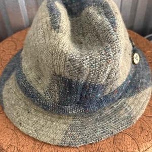 May include: A textured fedora hat in shades of olive green and blue. The hat features a rounded crown and a brim with a darker blue band. A small button detail is visible on the side. The hat is made of a woven material.