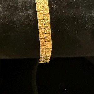 May include: A gold-toned bracelet featuring two parallel rows of square-shaped links. Each individual link has a textured, hammered surface that reflects light, creating a faceted appearance across the entire length of the jewelry piece.