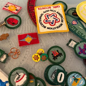 Op de afbeelding: Een verzameling Girl Scout-patches en -spelden. Patches zijn onder meer een rood, wit en blauw Bicentennial-patch en diverse groene en witte ronde patches met geborduurde ontwerpen. Sommige spelden zijn stervormig.