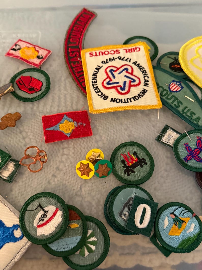Op de afbeelding: Een verzameling Girl Scout-patches en -spelden. Patches zijn onder meer een rood, wit en blauw Bicentennial-patch en diverse groene en witte ronde patches met geborduurde ontwerpen. Sommige spelden zijn stervormig.