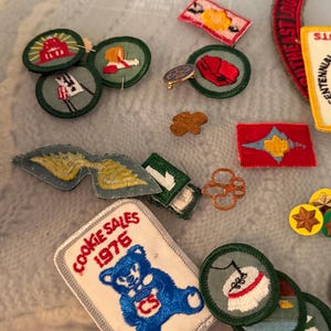 Op de afbeelding: Een verzameling vintage Girl Scout-patches en -spelden. De patches tonen verschillende ontwerpen, waaronder een beer met de tekst "Cookie Sales 1976", vlaggen en andere emblemen. De spelden omvatten een goudkleurige klaver en andere kleine badges.