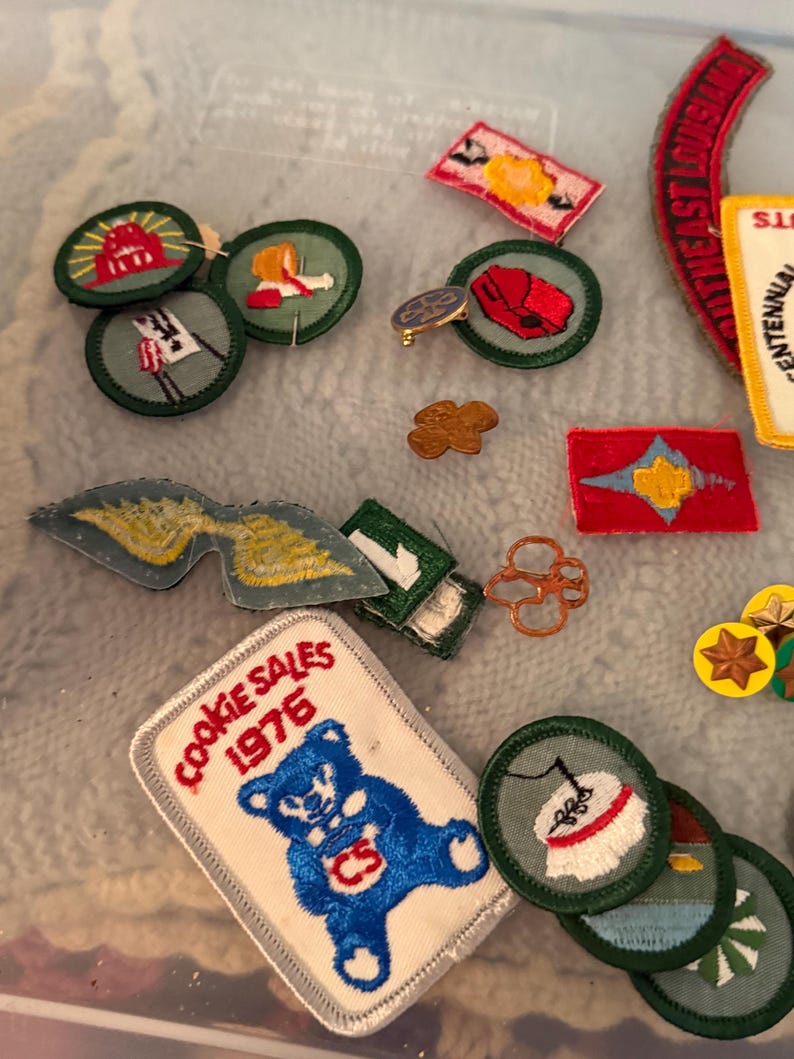 Op de afbeelding: Een verzameling vintage Girl Scout-patches en -spelden. De patches tonen verschillende ontwerpen, waaronder een beer met de tekst "Cookie Sales 1976", vlaggen en andere emblemen. De spelden omvatten een goudkleurige klaver en andere kleine badges.