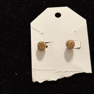 May include: A pair of gold-toned spherical stud earrings featuring a textured surface covered in small, sparkling crystals. The earrings are mounted on a white rectangular display card with a hole at the top for hanging and horizontal slits.