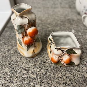 May include: A ceramic creamer pitcher and a matching sugar bowl featuring a cream-colored glaze with hand-painted orange fruit and dark green leaf motifs. Both pieces have a textured, organic shape with raised relief details and gold trim.