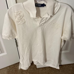 May include: A white short-sleeved polo shirt by Polo by Ralph Lauren featuring a large embroidered number 2 on the right chest and tonal embroidery on the left chest. The shirt has a classic collar, a buttoned placket, and a vented hemline.