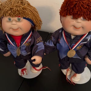 May include: Two Cabbage Patch Kids dolls wearing blue and navy tracksuits with gold medals. One doll has brown yarn hair and a blue headband, the other has red yarn hair. Both dolls are on white platforms.
