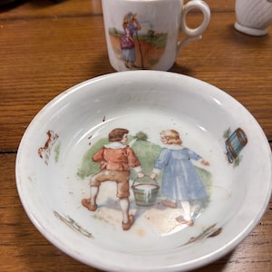 May include: A small, white ceramic cup and saucer set. The cup has a handle and a painted scene of a person in a field. The saucer has a painted scene of two children carrying a bucket. The set is antique.