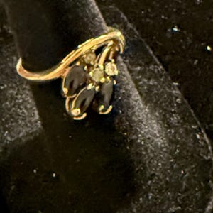 May include: A gold ring featuring a cluster of dark, oval-shaped stones arranged in a floral pattern, accented by small, clear, round stones set within the center of the arrangement. The band is thin and smooth, curving slightly to support the setting.