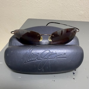 May include: Rimless Maui Jim sunglasses with dark brown gradient lenses and thin bronze-colored metal temples. The sunglasses rest on a black, oval-shaped hard protective case featuring the embossed Maui Jim logo on the front surface.